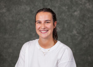 A smiling individual with long hair, wearing a simple shirt and layered necklaces, poses against a textured backdrop.